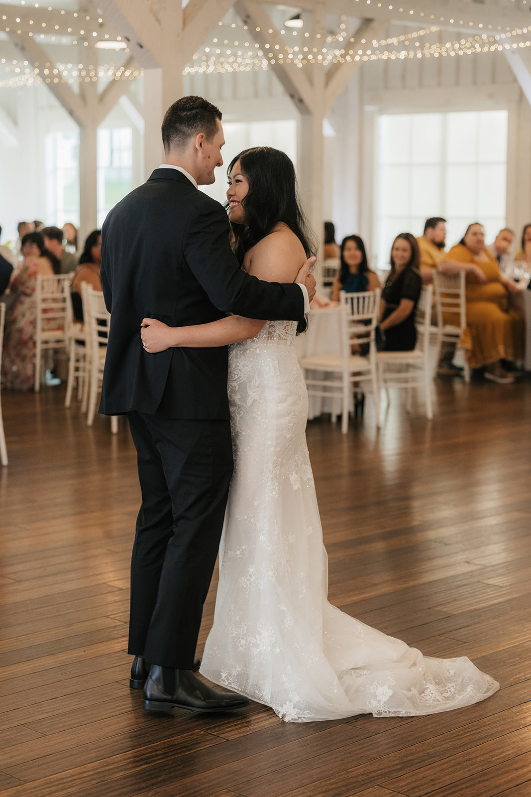 A bride and groom share their first dance in a bright, decorated reception hall with seated guests watching in the background.