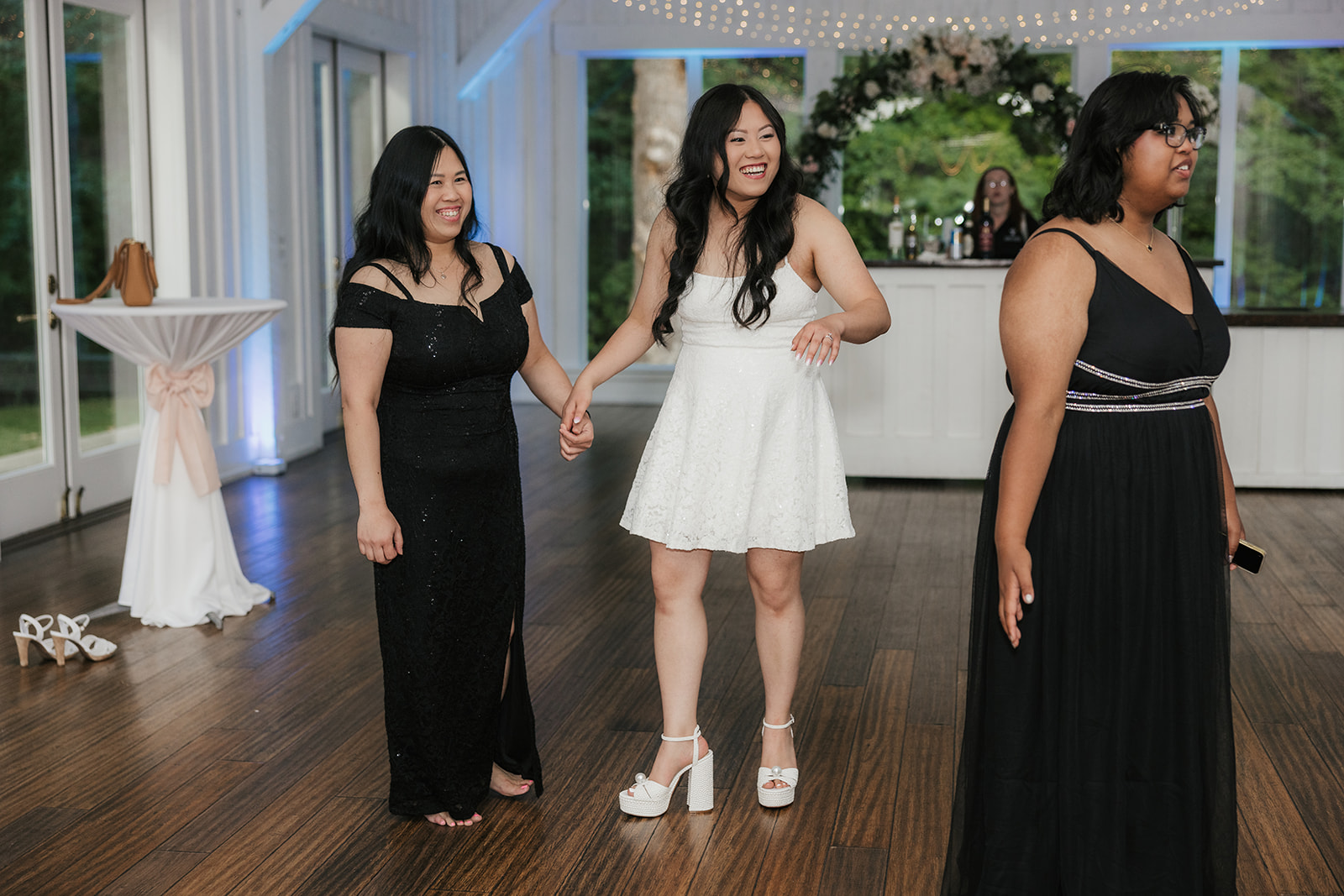 A group of women in formal dresses stand and talk on a wooden floor in a decorated indoor venue with string lights overhead.