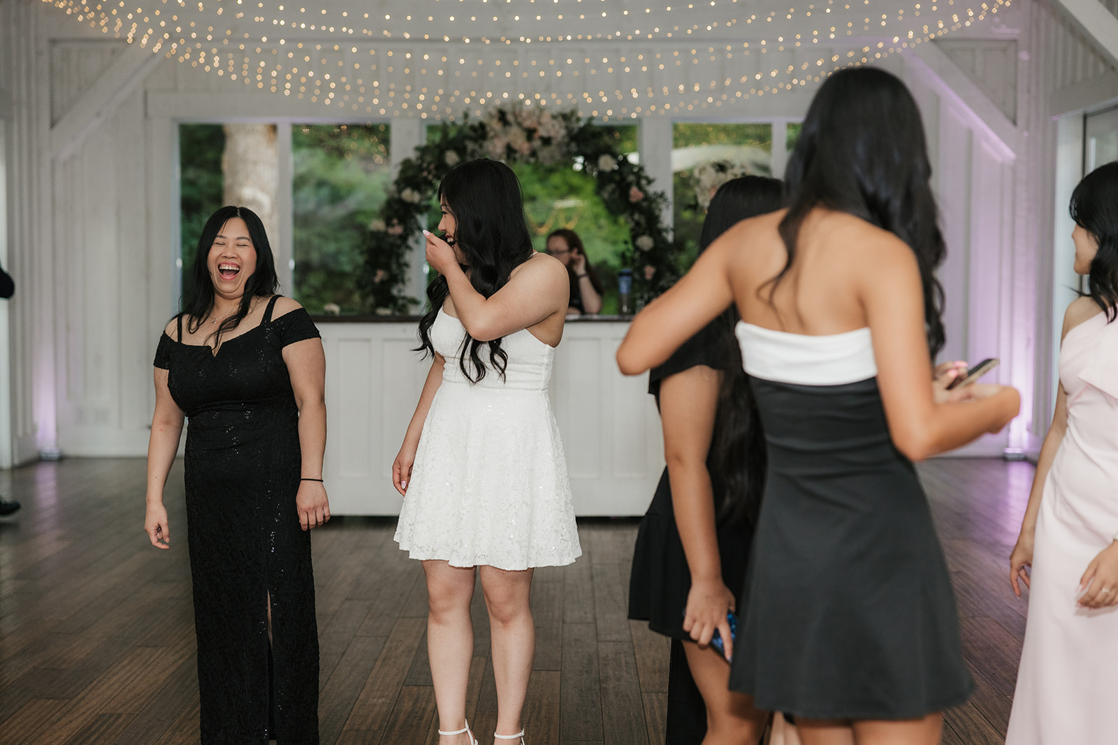 A group of women in formal dresses stand and talk on a wooden floor in a decorated indoor venue with string lights overhead.