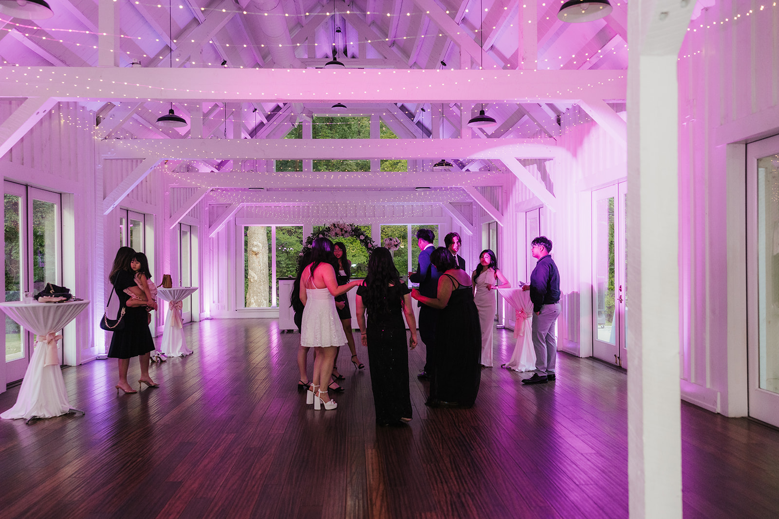 A group of people dressed in formal attire stand and talk in a spacious, well-lit event hall with wooden floors and purple accent lighting.
