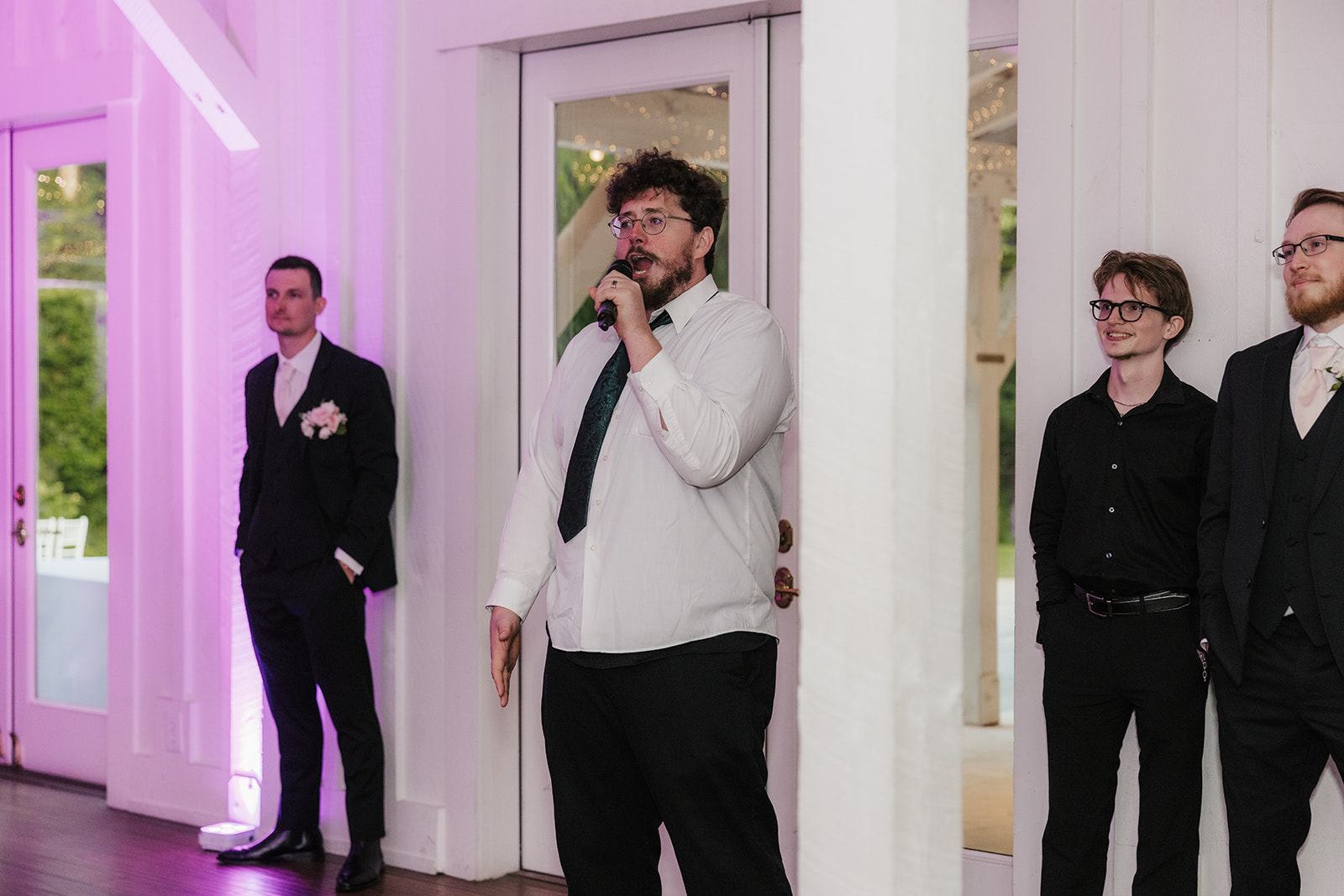 A group of people dressed in formal attire stand and talk in a spacious, well-lit event hall with wooden floors and purple accent lighting.