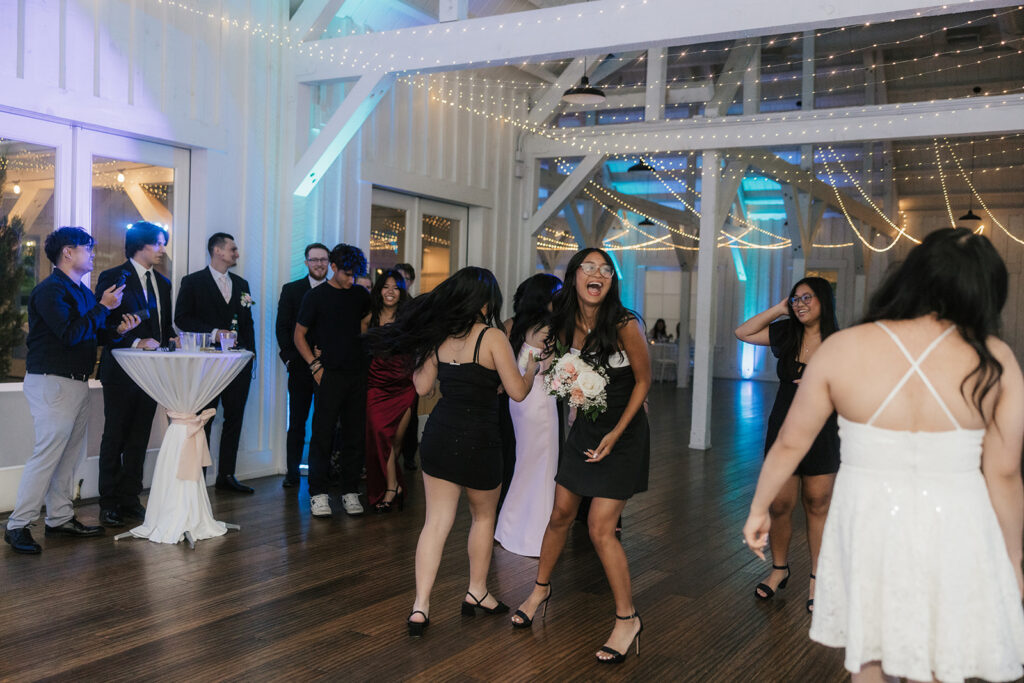 A group of people dressed in formal attire stand and talk in a spacious, well-lit event hall with wooden floors and purple accent lighting.