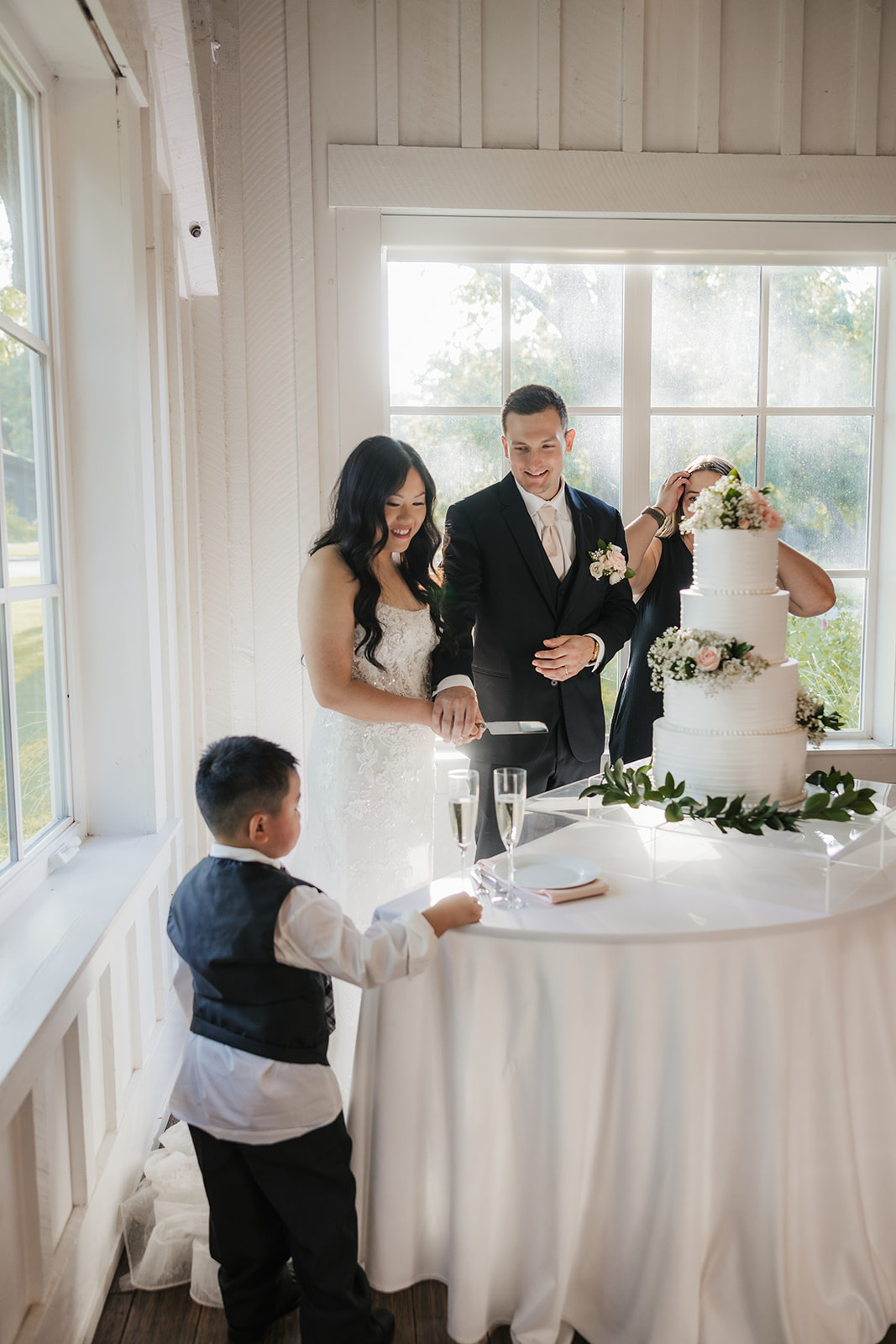 A bride and groom stand together cutting a three-tiered white wedding cake decorated with flowers, near a window with natural light.