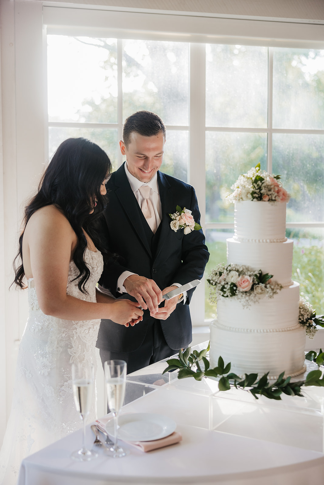 A bride and groom stand together cutting a three-tiered white wedding cake decorated with flowers, near a window with natural light.