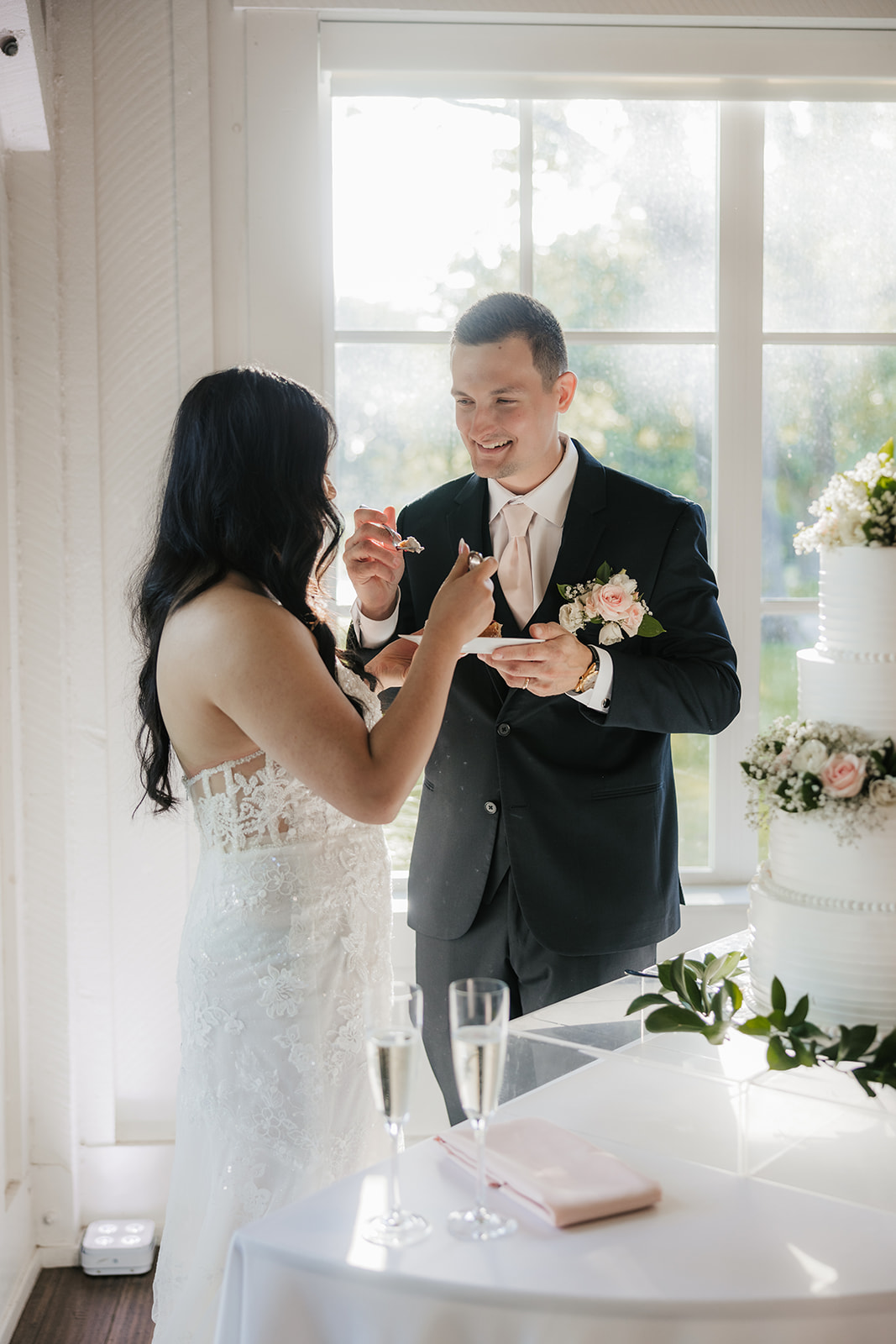 A bride and groom stand together cutting a three-tiered white wedding cake decorated with flowers, near a window with natural light.