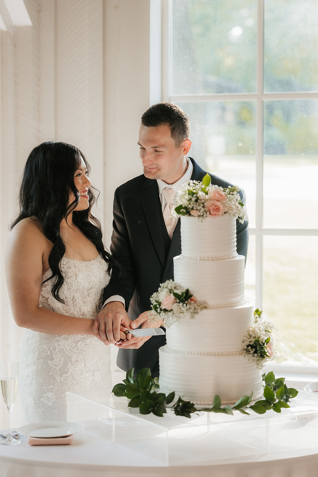 A bride and groom stand together cutting a three-tiered white wedding cake decorated with flowers, near a window with natural light.