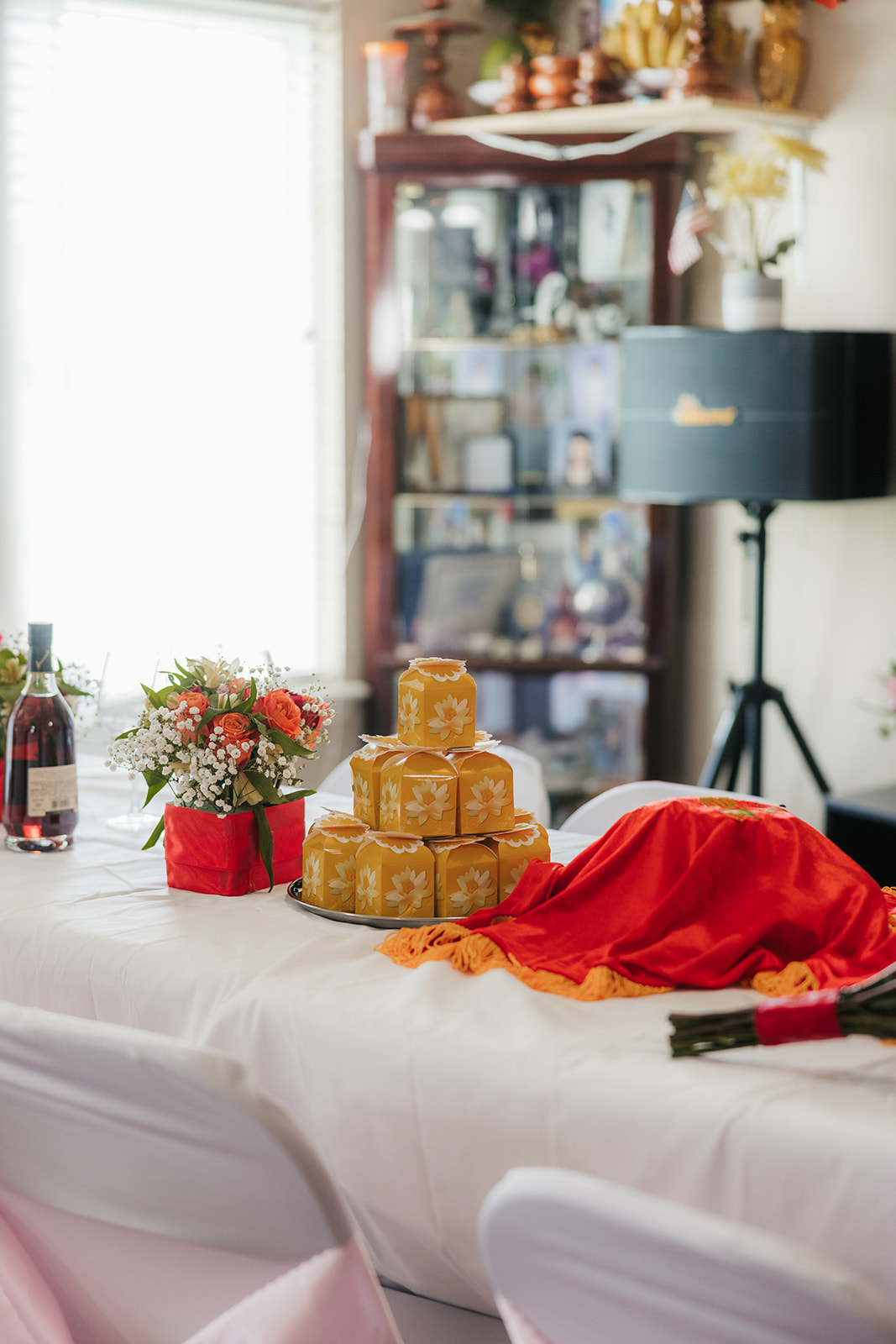 A table with a stack of golden boxes, a red cloth, wine bottle, floral arrangement, and decorative items, set in a brightly lit room.