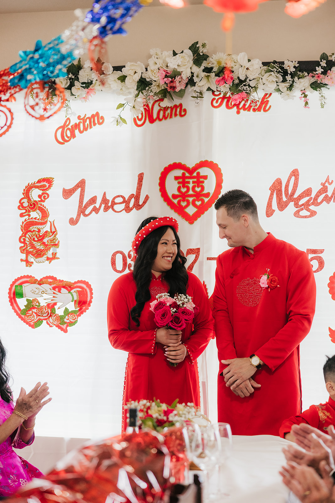 A couple dressed in traditional red attire stands smiling at a Vietnamese engagement ceremony, with decorated backdrop and guests seated at a table in the foreground.