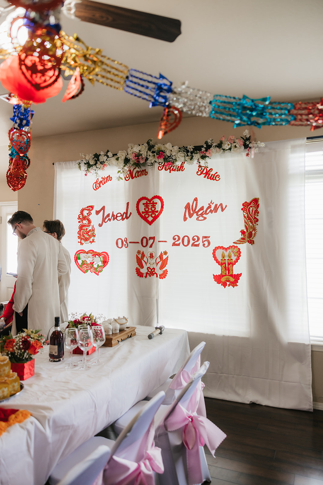 A table with a stack of golden boxes, a red cloth, wine bottle, floral arrangement, and decorative items, set in a brightly lit room.