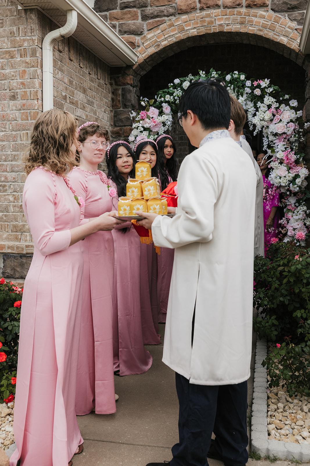 A group of women in pink dresses face a man in a white outfit, holding a tray with yellow items, standing outside a brick building decorated with flowers.