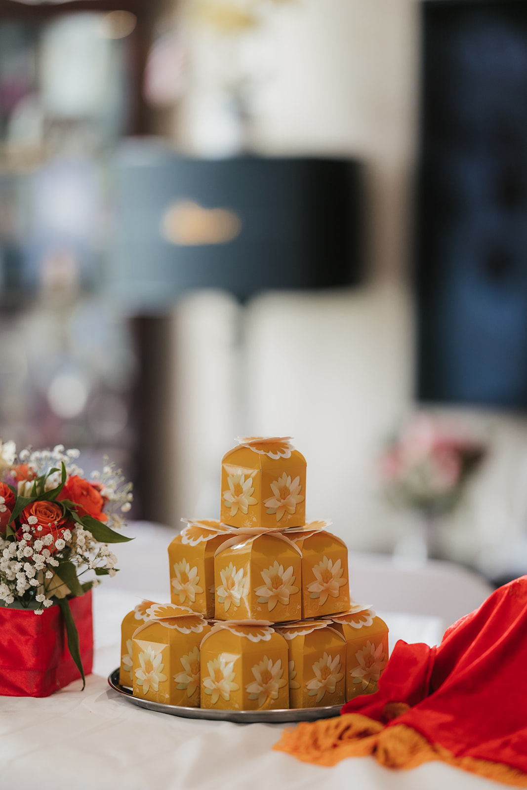 A stack of square yellow sweets with white designs is arranged on a round tray, with a floral arrangement and red cloth nearby.