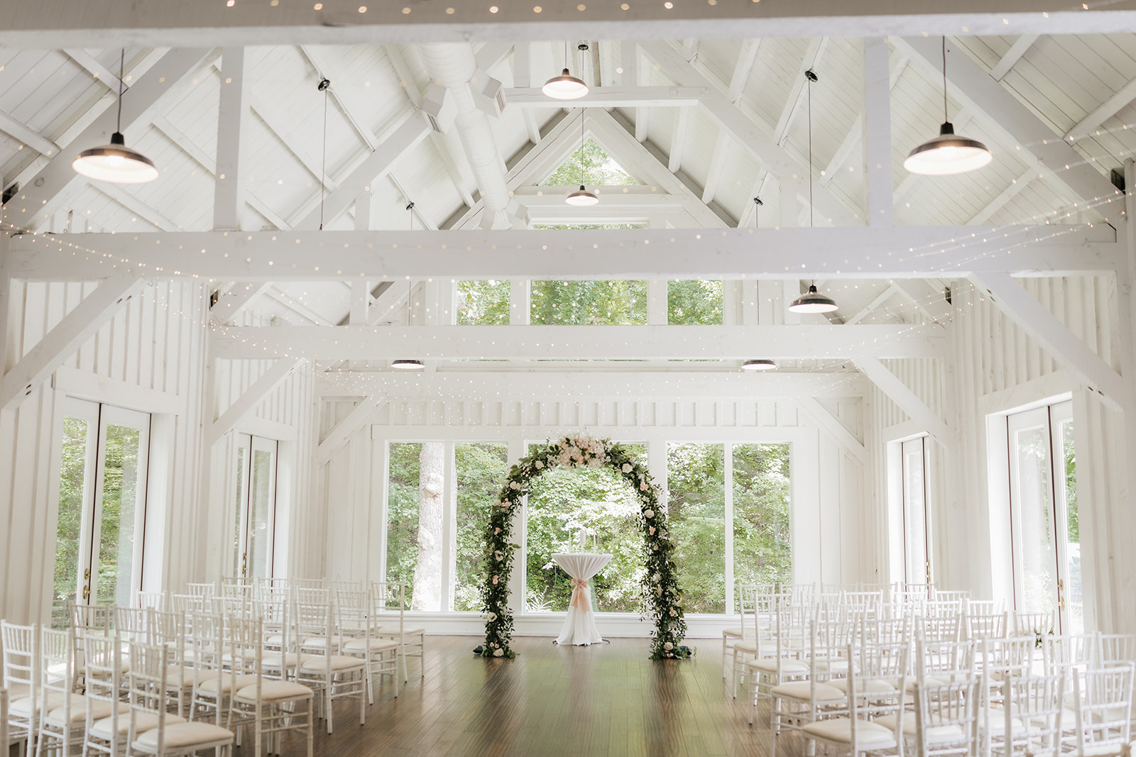 Bright, white wedding venue with high ceilings, large windows, rows of white chairs, and a floral arch at the center of the room.