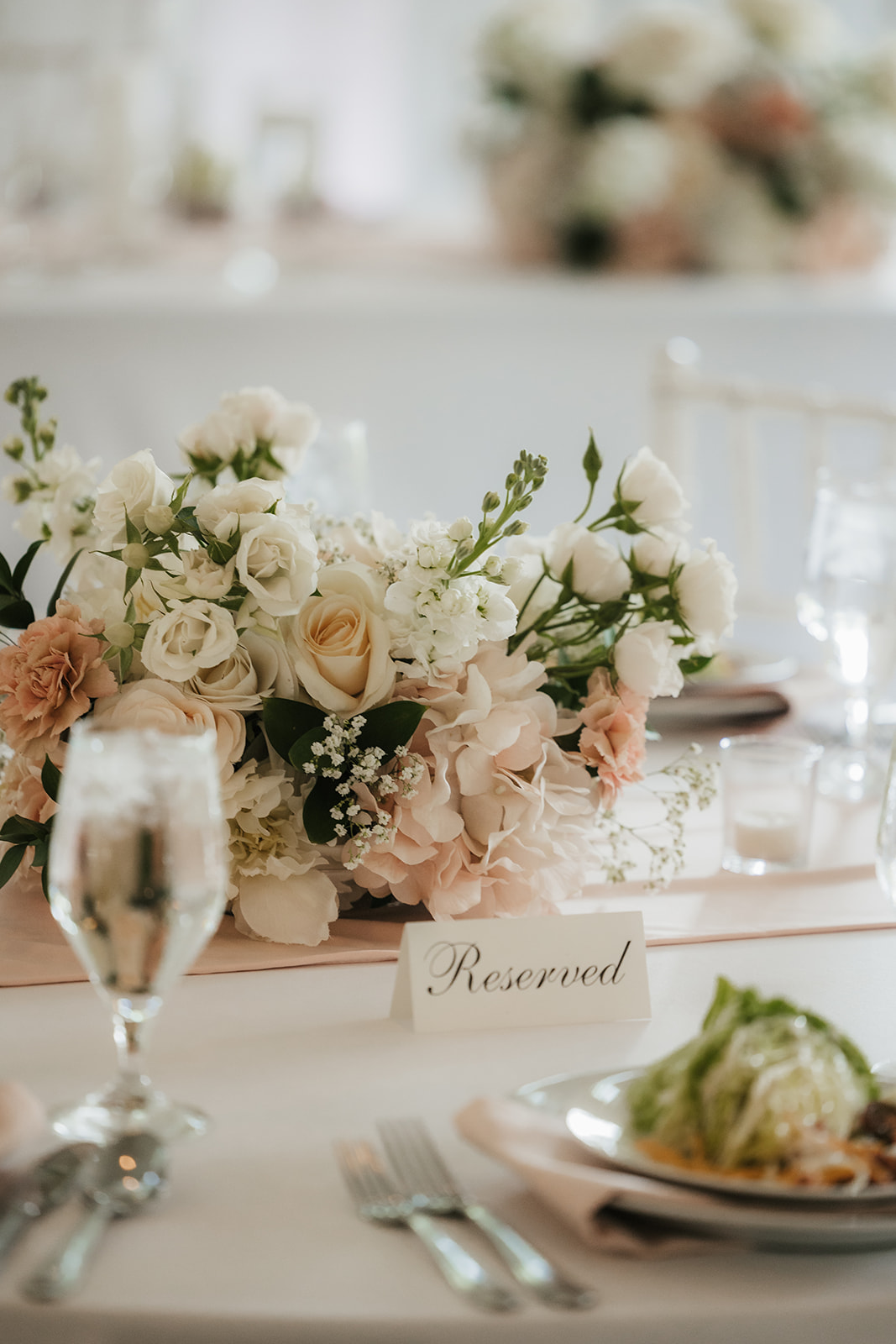 A bright, airy event space with white walls and wooden beams, featuring round tables set with white linens and floral centerpieces, arranged for a celebration for a wedding at spain ranch