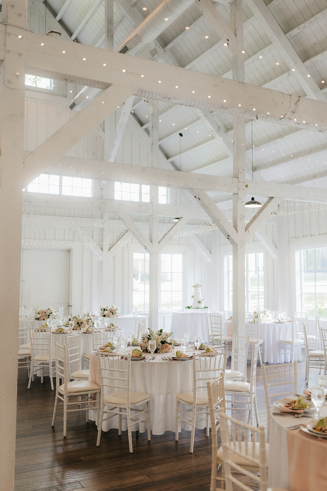 A bright, airy event space with white walls and wooden beams, featuring round tables set with white linens and floral centerpieces, arranged for a celebration for a wedding at spain ranch