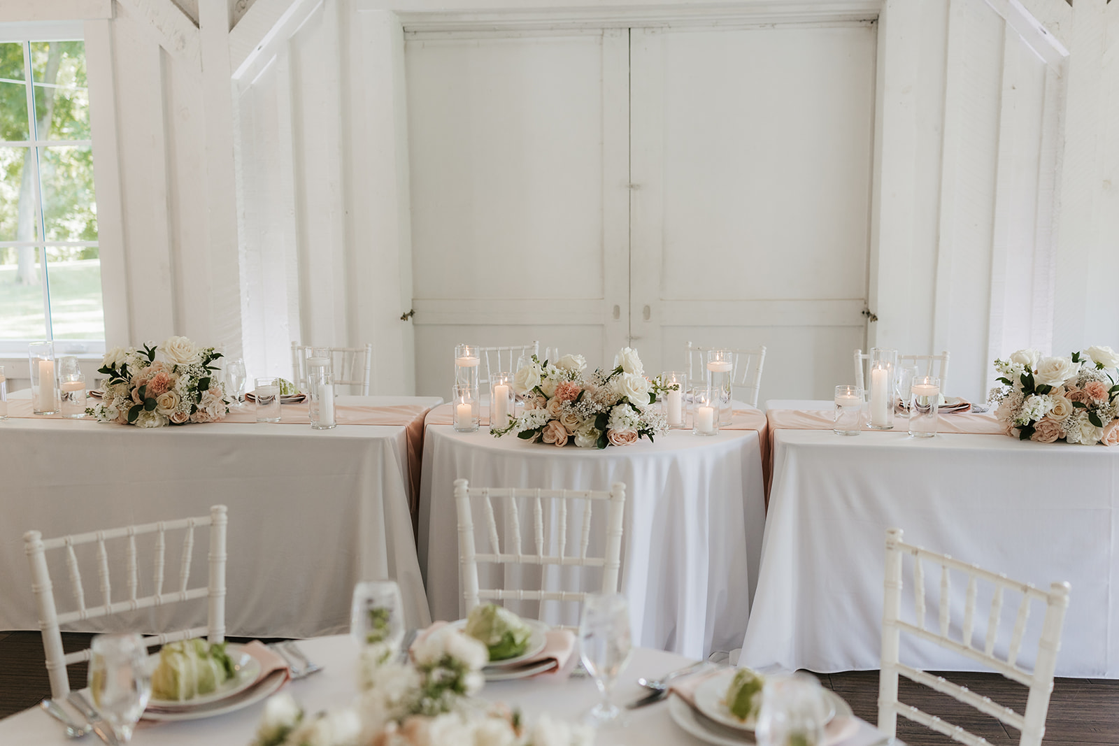 A bright, airy event space with white walls and wooden beams, featuring round tables set with white linens and floral centerpieces, arranged for a celebration for a wedding at spain ranch