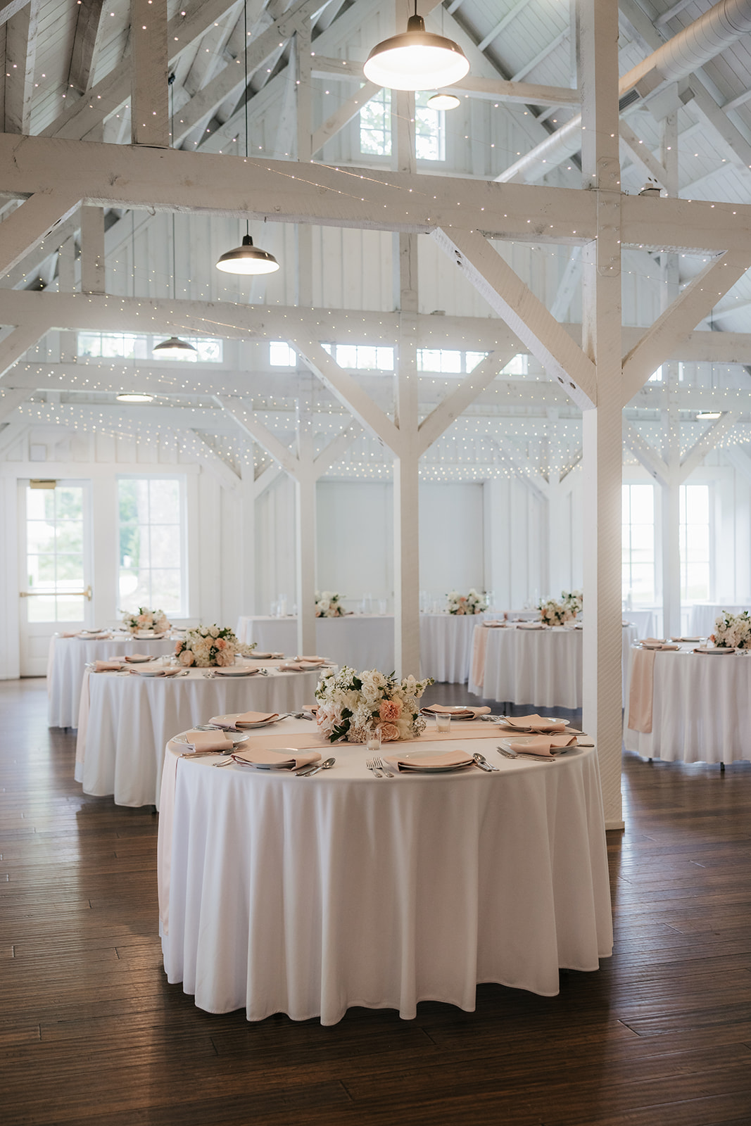 A bright, airy event space with white walls and wooden beams, featuring round tables set with white linens and floral centerpieces, arranged for a celebration for a wedding at spain ranch