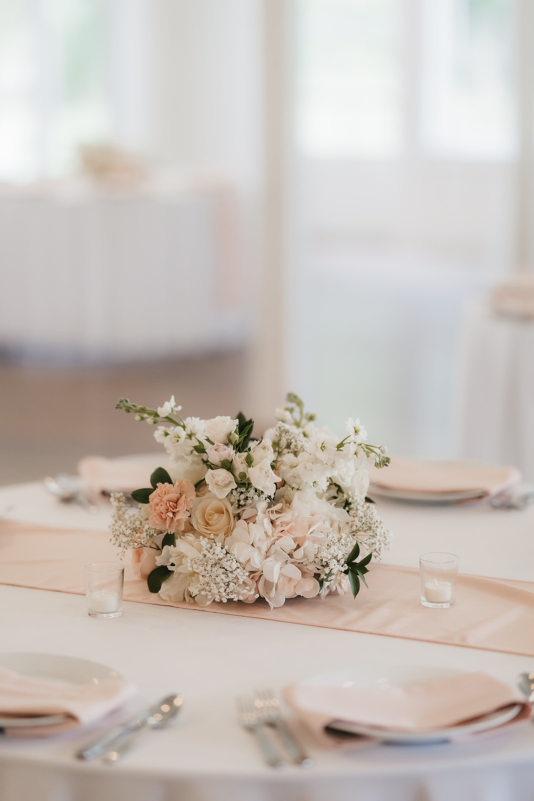 A round table set for a meal with plates, cutlery, blush napkins, and a centerpiece of white and pale pink flowers in a bright, softly lit room.