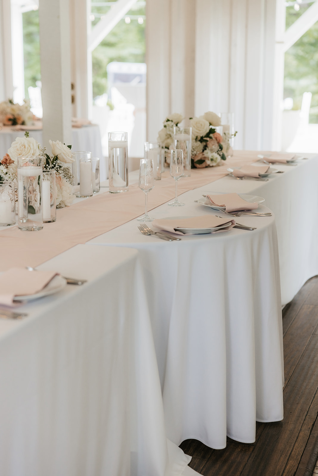 A bright, airy event space with white walls and wooden beams, featuring round tables set with white linens and floral centerpieces, arranged for a celebration for a wedding at spain ranch