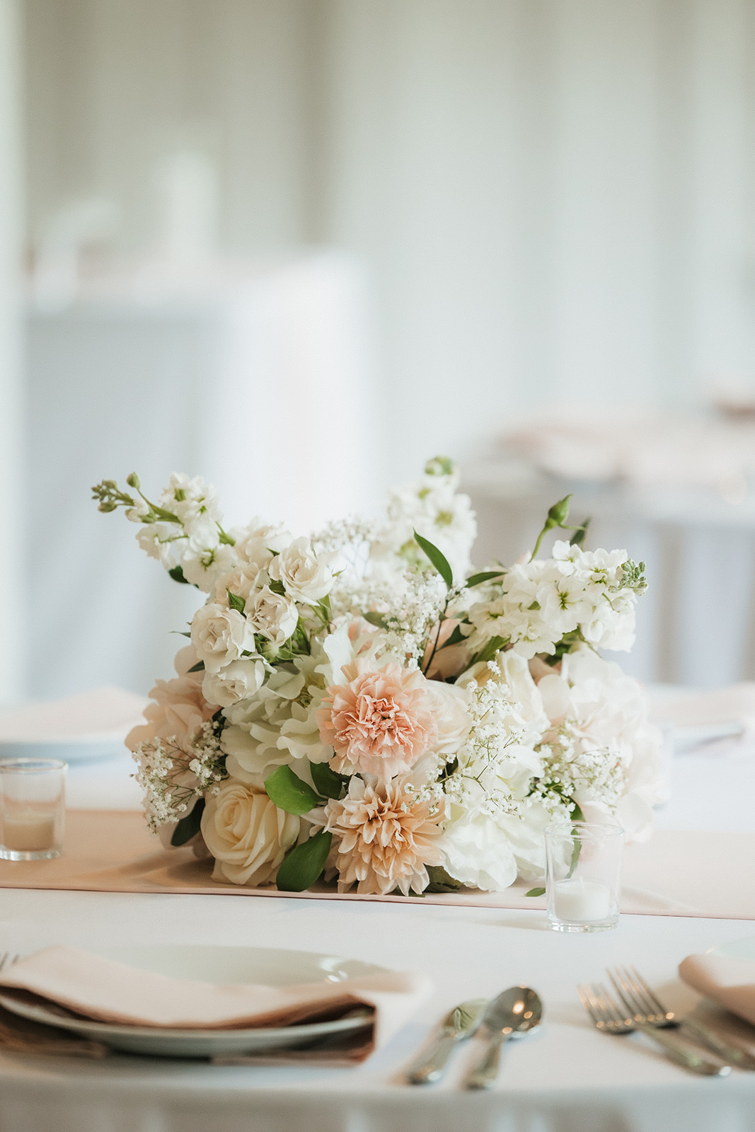 A bright, airy event space with white walls and wooden beams, featuring round tables set with white linens and floral centerpieces, arranged for a celebration for a wedding at spain ranch