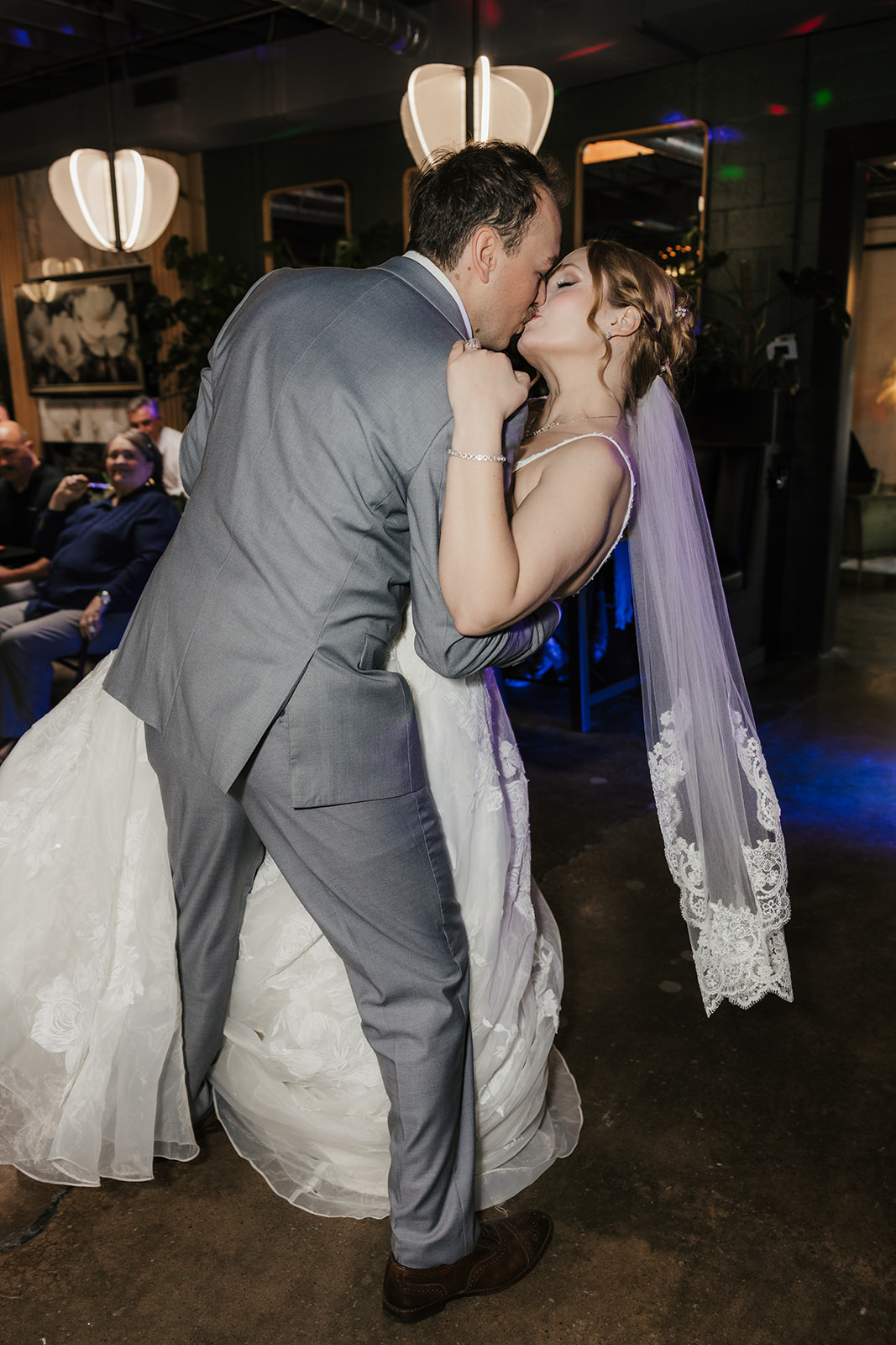 A bride and groom in formal wedding attire share a dance on a dimly lit dance floor, with guests seated in the background at a Camp Loughridge Wedding