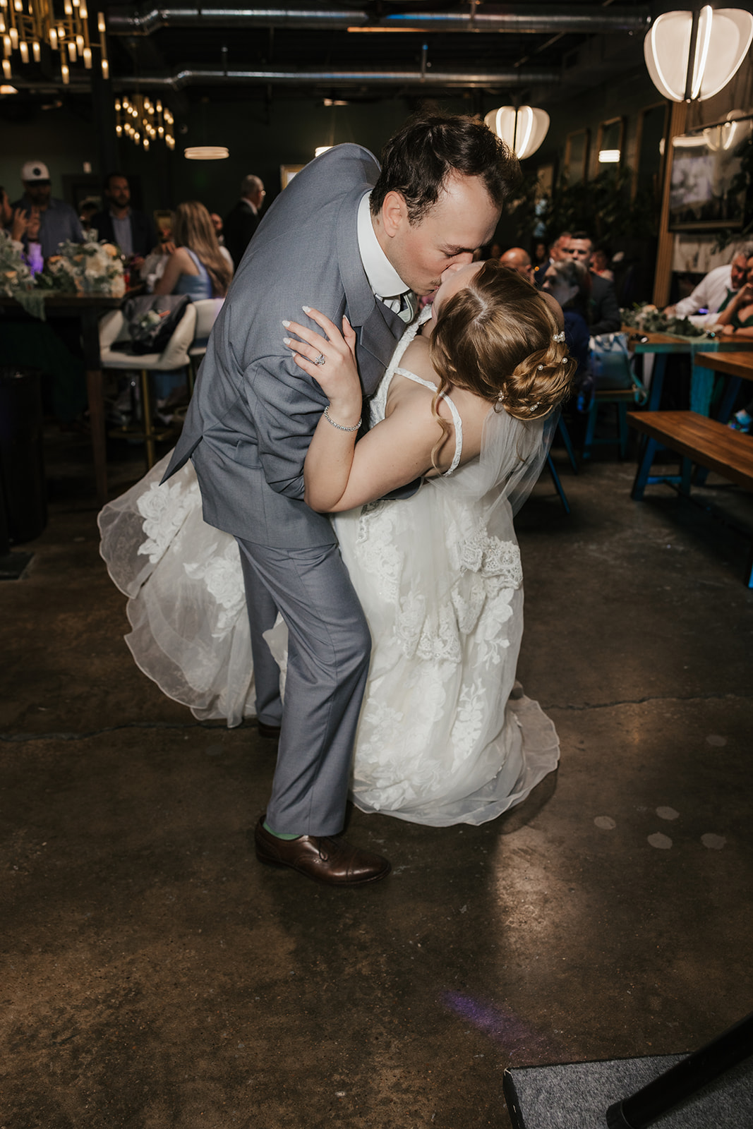 A bride and groom in formal wedding attire share a dance on a dimly lit dance floor, with guests seated in the background at a Camp Loughridge Wedding