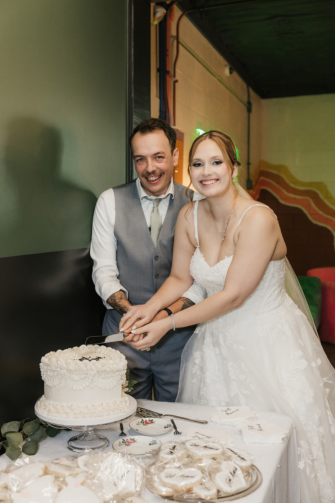 A bride and groom stand together, smiling, as they cut a white wedding cake at a decorated table at a Camp Loughridge Wedding
