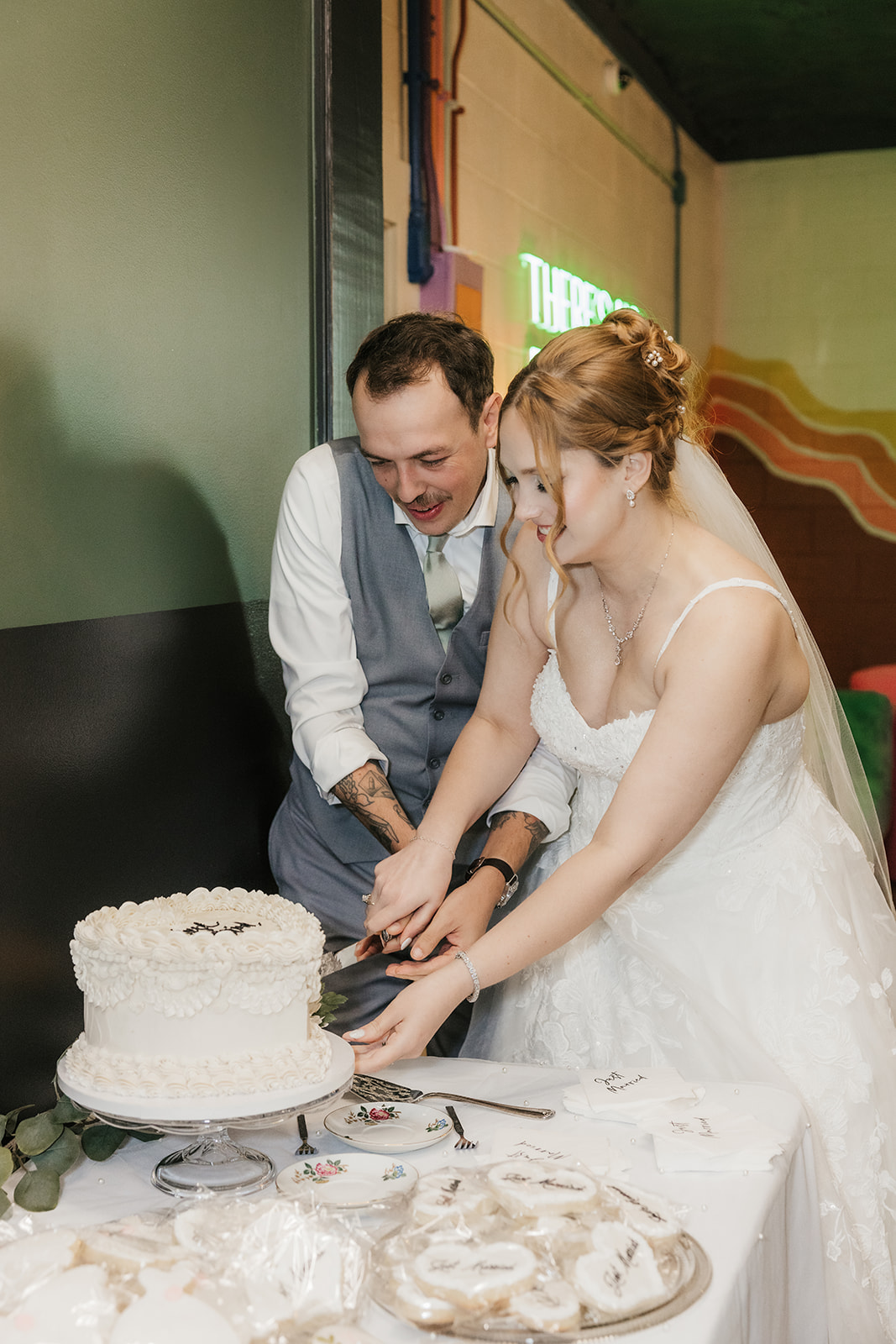 A bride and groom stand together, smiling, as they cut a white wedding cake at a decorated table at a Camp Loughridge Wedding