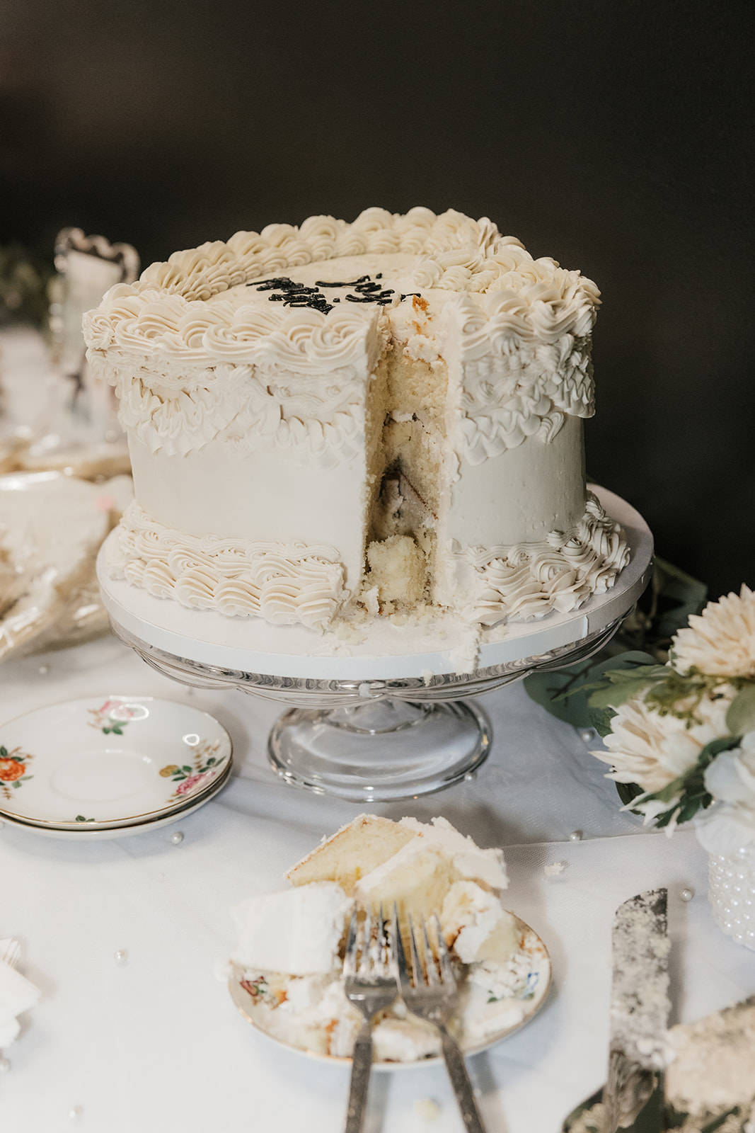 A round white frosted cake with a slice removed sits on a cake stand, with a plate containing a piece of the cake and utensils nearby at a Camp Loughridge Wedding