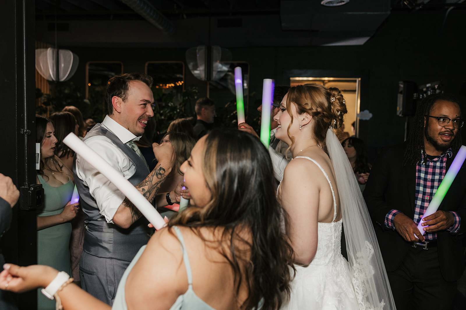 A bride and guests, holding light-up sticks, dance and celebrate together at an indoor wedding reception for a wedding at Camp Loughridge Wedding