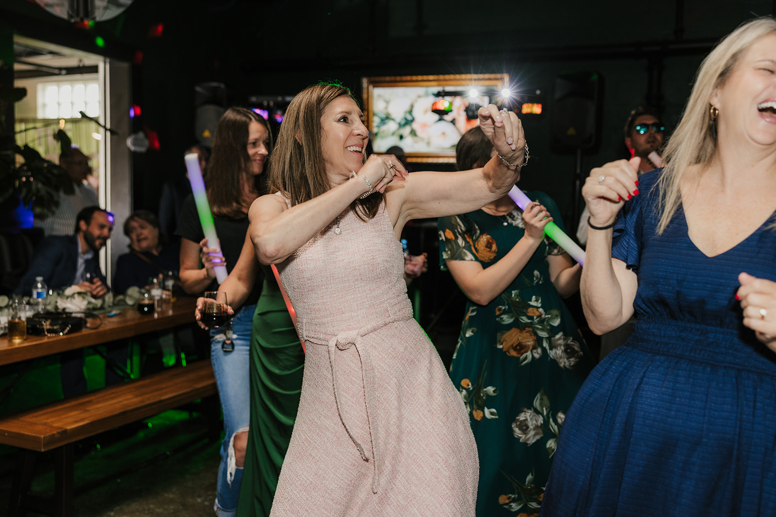 A bride and guests, holding light-up sticks, dance and celebrate together at an indoor wedding reception for a wedding at Camp Loughridge Wedding
