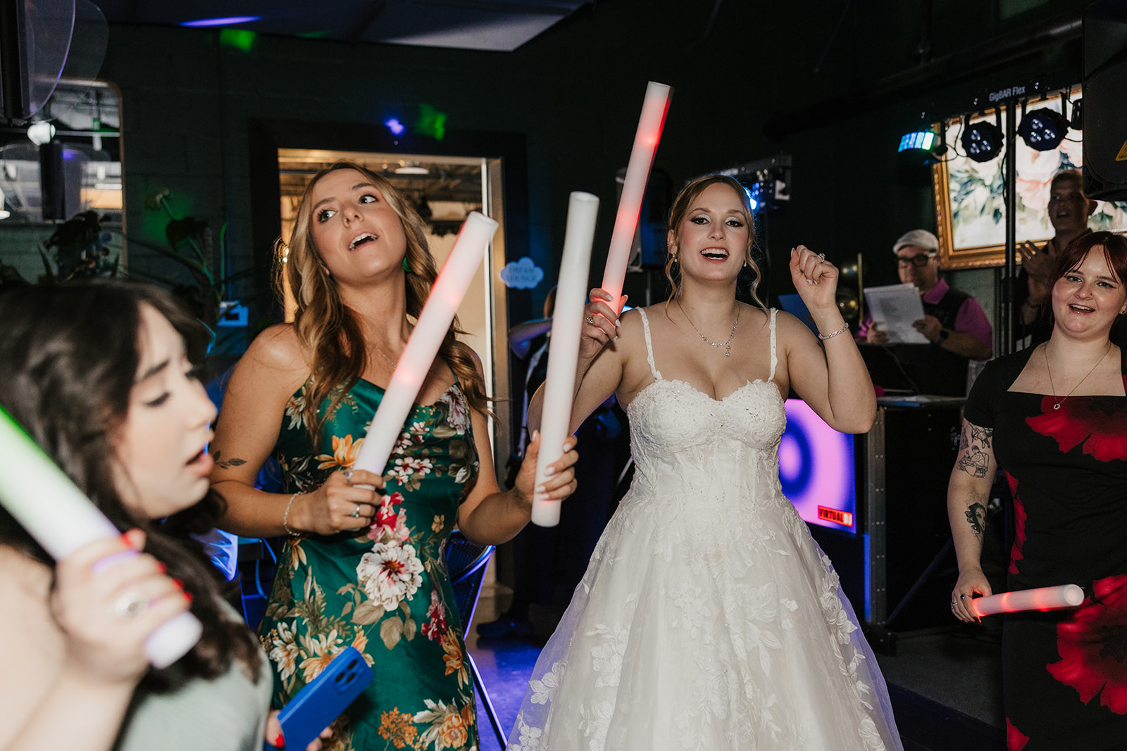 A bride and guests, holding light-up sticks, dance and celebrate together at an indoor wedding reception for a wedding at Camp Loughridge Wedding
