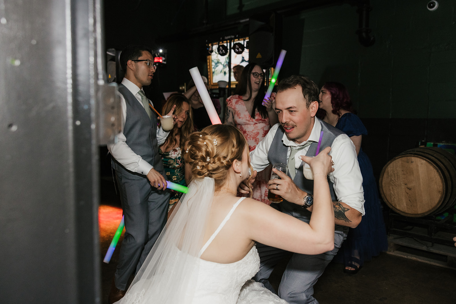 A bride and guests, holding light-up sticks, dance and celebrate together at an indoor wedding reception for a wedding at Camp Loughridge Wedding