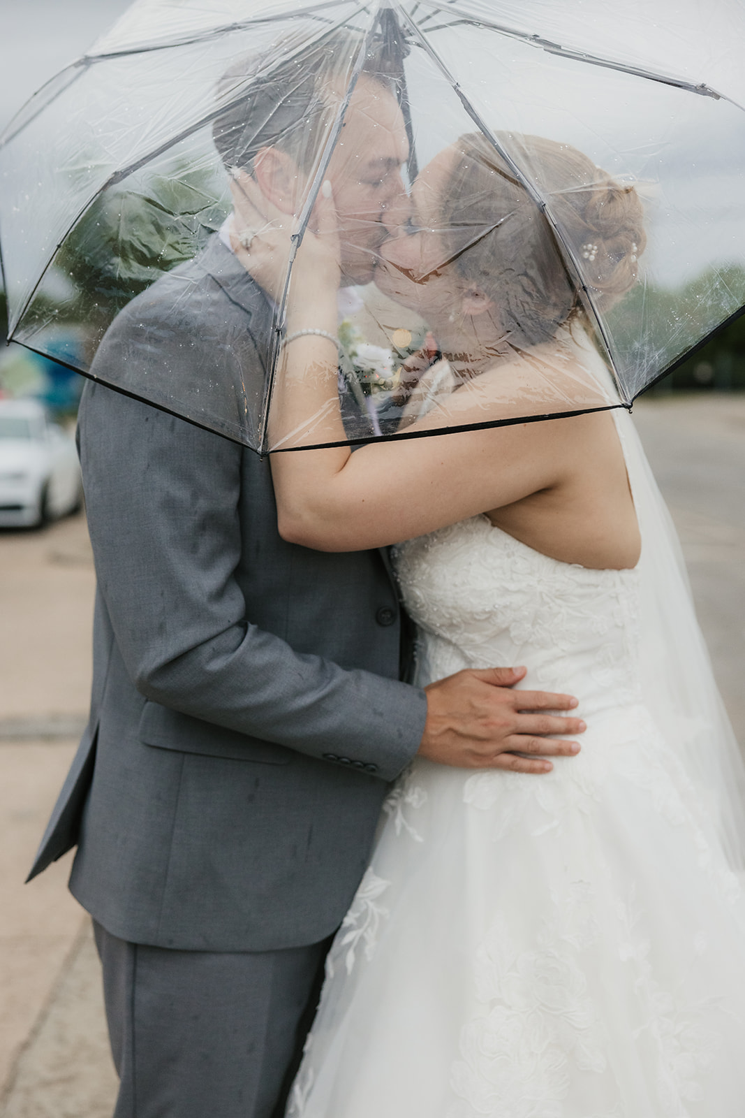 A bride and groom stand together under a clear umbrella on a city sidewalk, with an overcast sky in the background at a Camp Loughridge Wedding