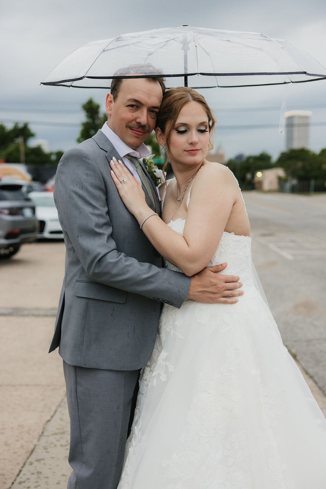 A bride and groom stand together under a clear umbrella on a city sidewalk, with an overcast sky in the background at a Camp Loughridge Wedding