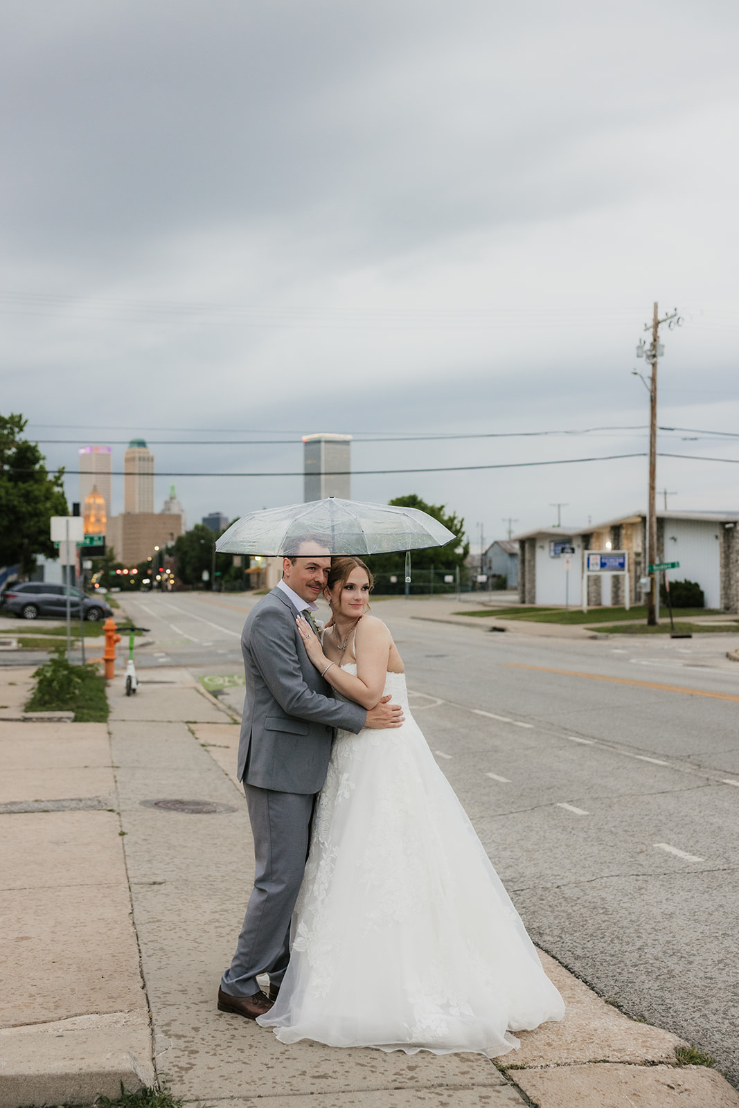 A bride and groom stand together under a clear umbrella on a city sidewalk, with an overcast sky in the background at a Camp Loughridge Wedding