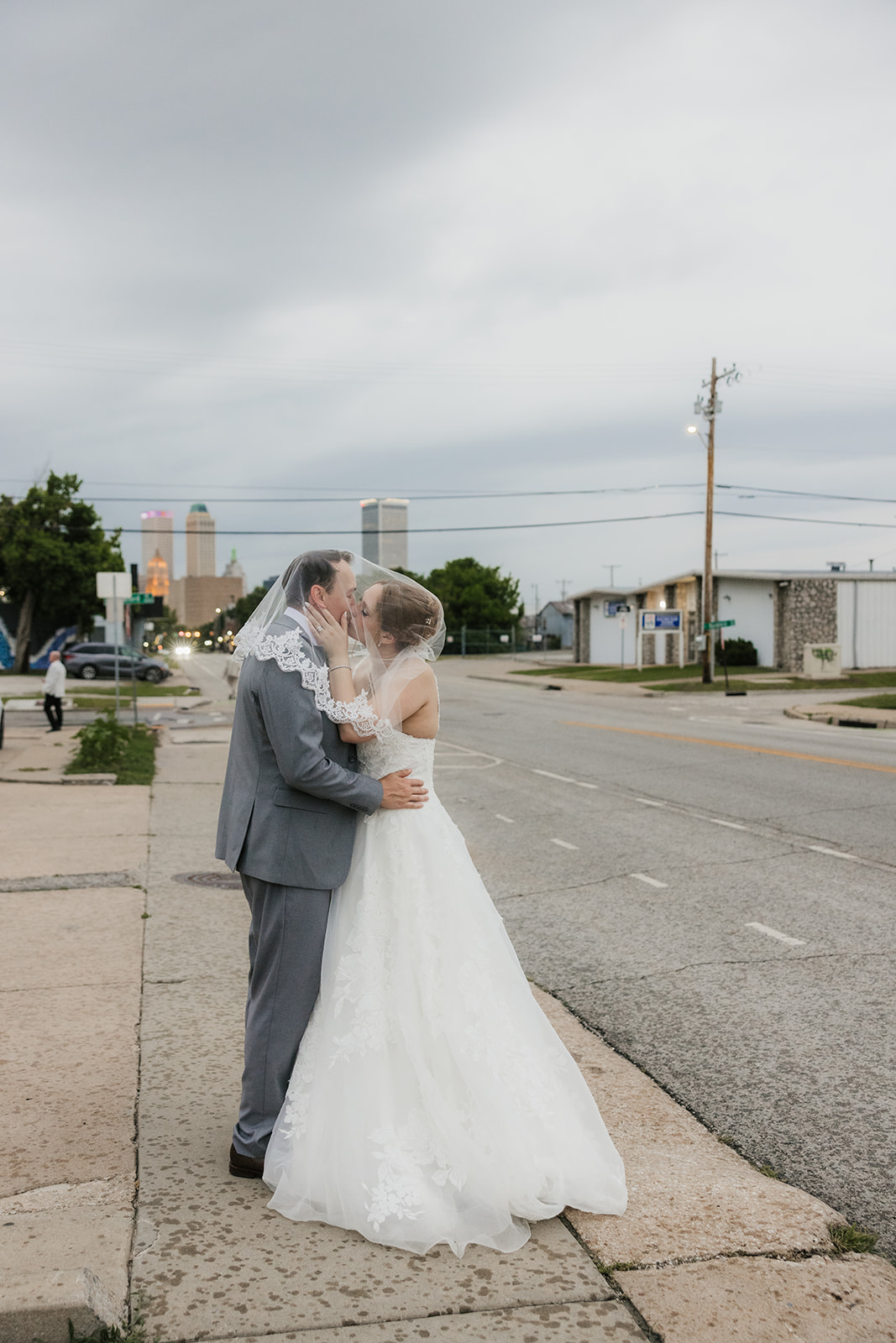 A bride and groom stand together under a clear umbrella on a city sidewalk, with an overcast sky in the background at a Camp Loughridge Wedding