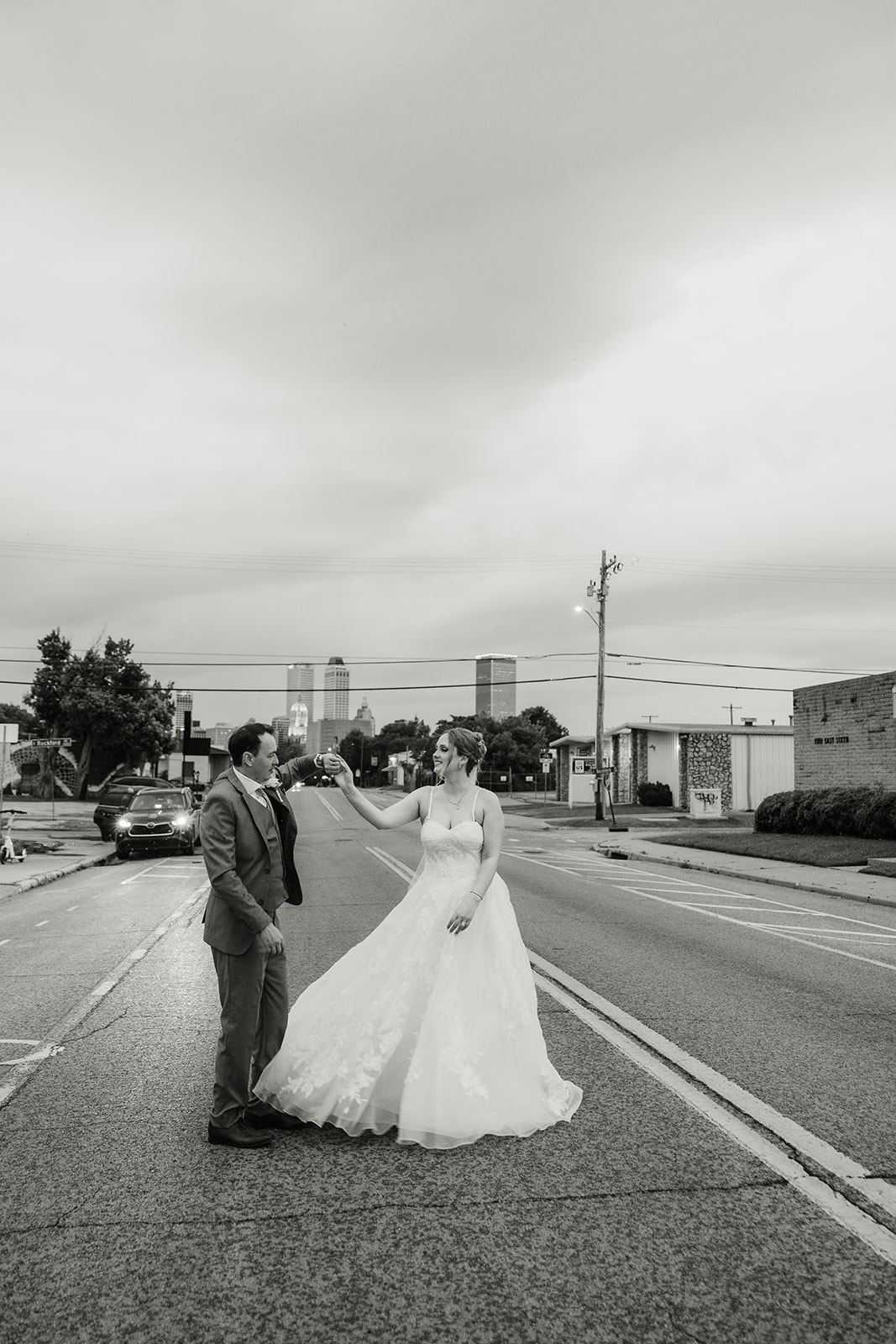A bride and groom stand together under a clear umbrella on a city sidewalk, with an overcast sky in the background at a Camp Loughridge Wedding