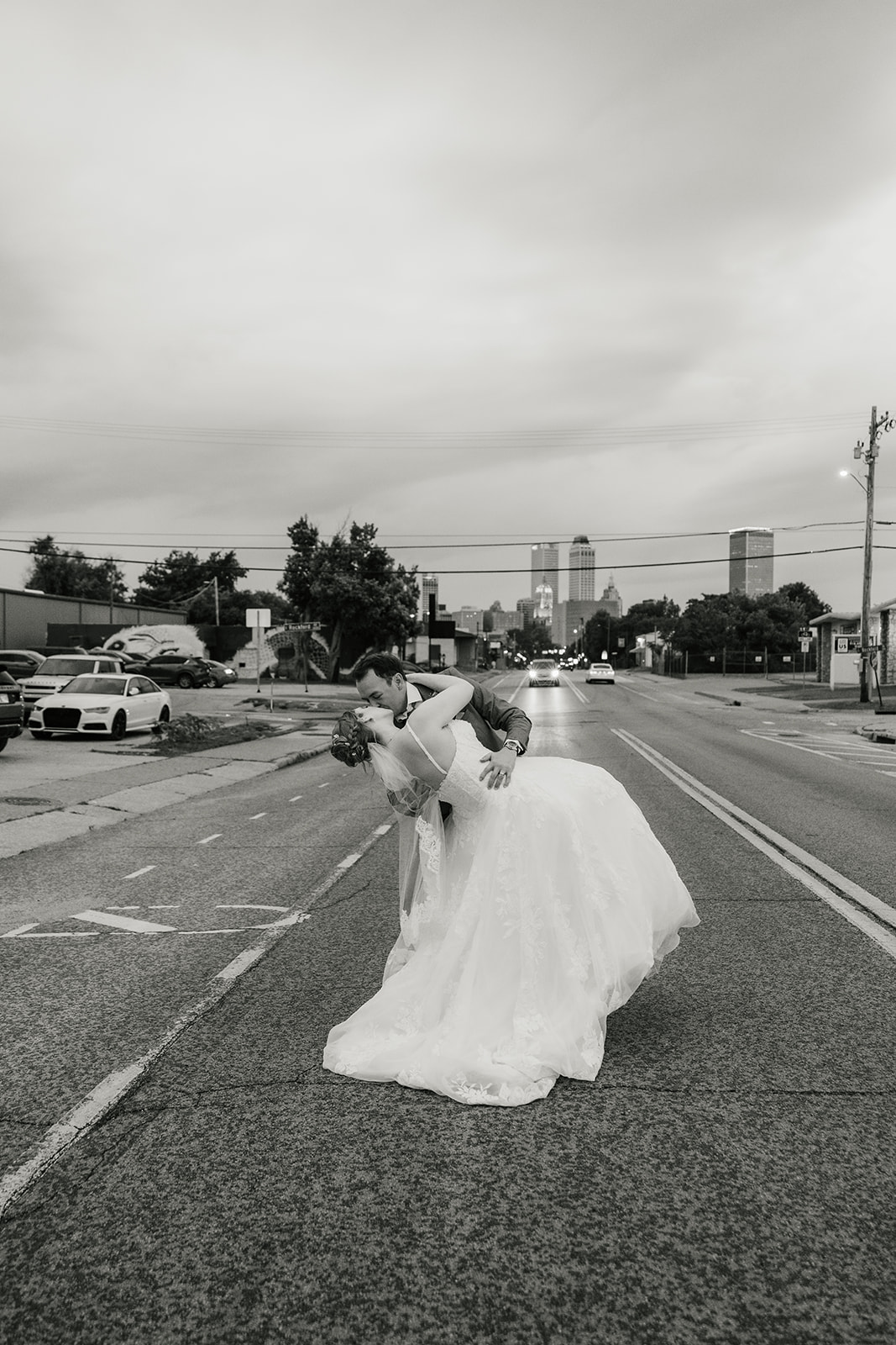 A bride and groom stand together under a clear umbrella on a city sidewalk, with an overcast sky in the background at a Camp Loughridge Wedding