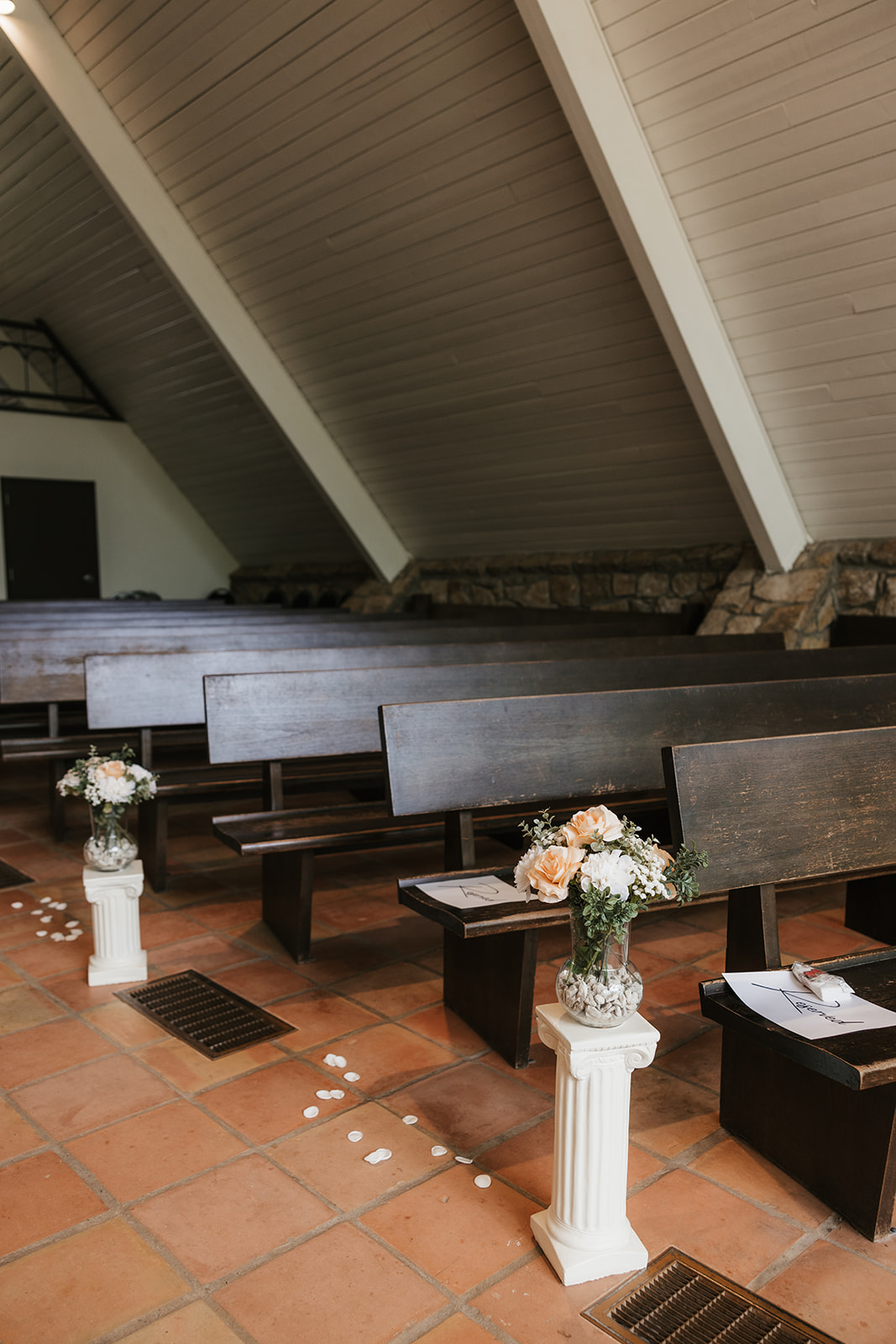 Wooden pews in an empty chapel, with flower arrangements on white pedestals and scattered flower petals lining the aisle.