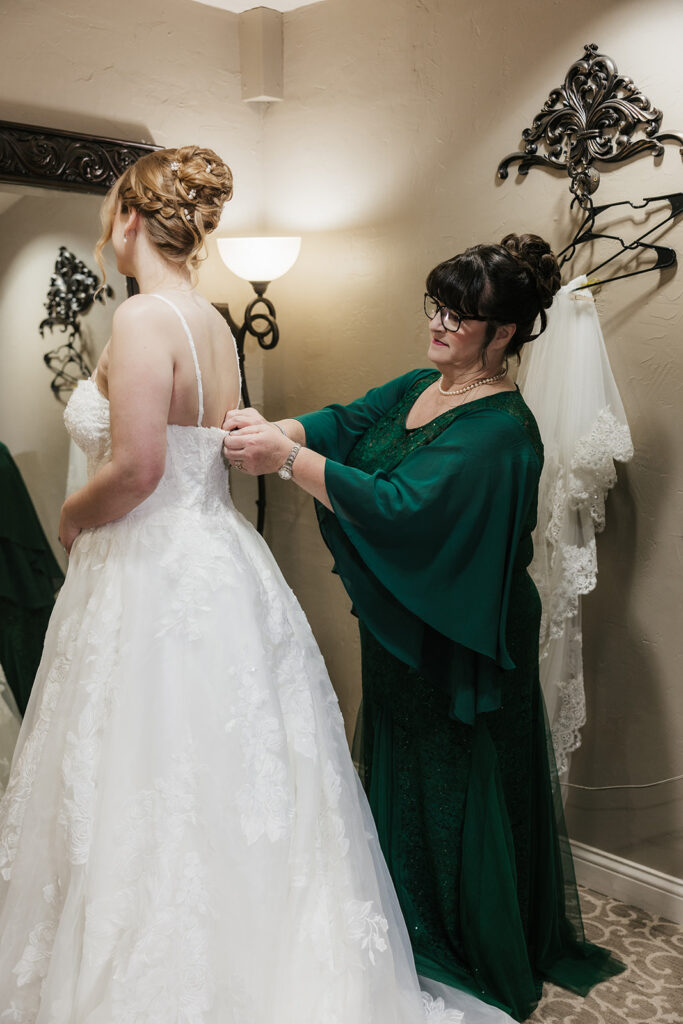 A woman in a green dress helps a bride adjust her white gown in a softly lit room with a wall mirror and decorative hooks.