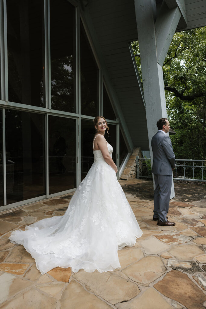 A bride in a white gown stands smiling while a groom in a gray suit faces away from her outside a modern glass building for a Camp Loughridge Wedding