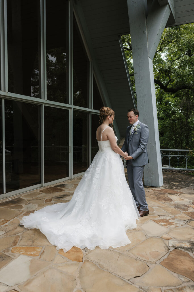 A bride in a white gown stands smiling while a groom in a gray suit faces away from her outside a modern glass building for a Camp Loughridge Wedding
