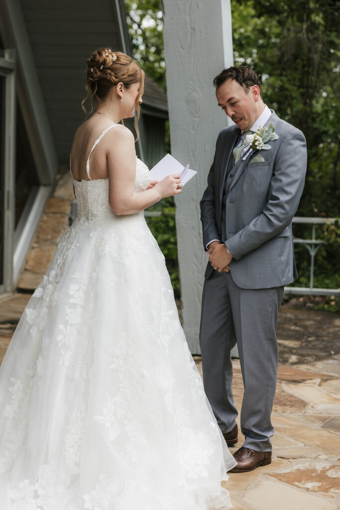 A bride in a white gown stands smiling while a groom in a gray suit faces away from her outside a modern glass building for a Camp Loughridge Wedding