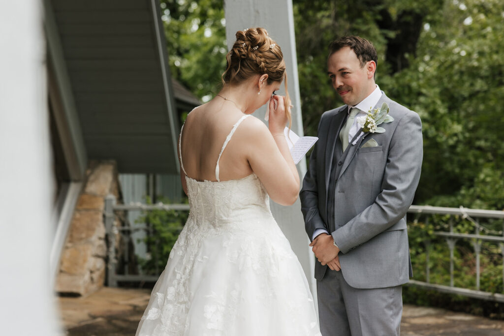 A bride in a white gown stands smiling while a groom in a gray suit faces away from her outside a modern glass building for a Camp Loughridge Wedding