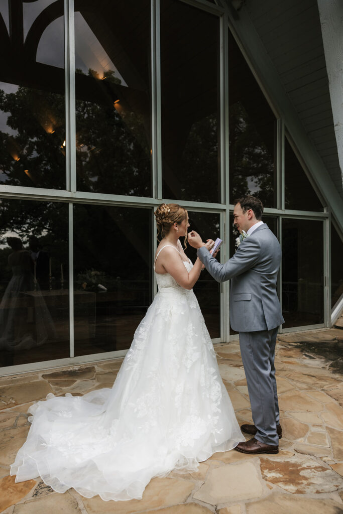 A bride in a white gown stands smiling while a groom in a gray suit faces away from her outside a modern glass building for a Camp Loughridge Wedding