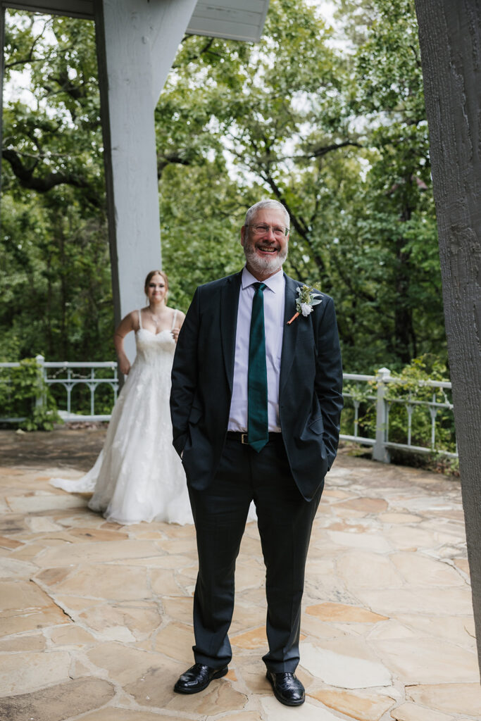 A bride in a white gown stands smiling while a groom in a gray suit faces away from her outside a modern glass building for a Camp Loughridge Wedding