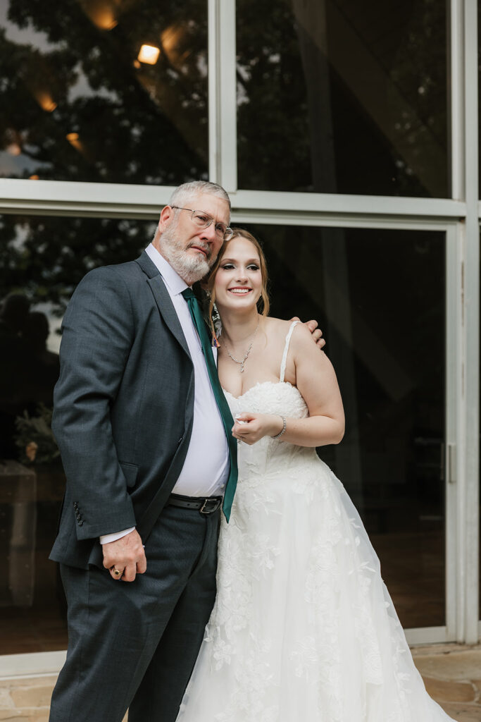 A bride in a white gown stands smiling while a groom in a gray suit faces away from her outside a modern glass building for a Camp Loughridge Wedding