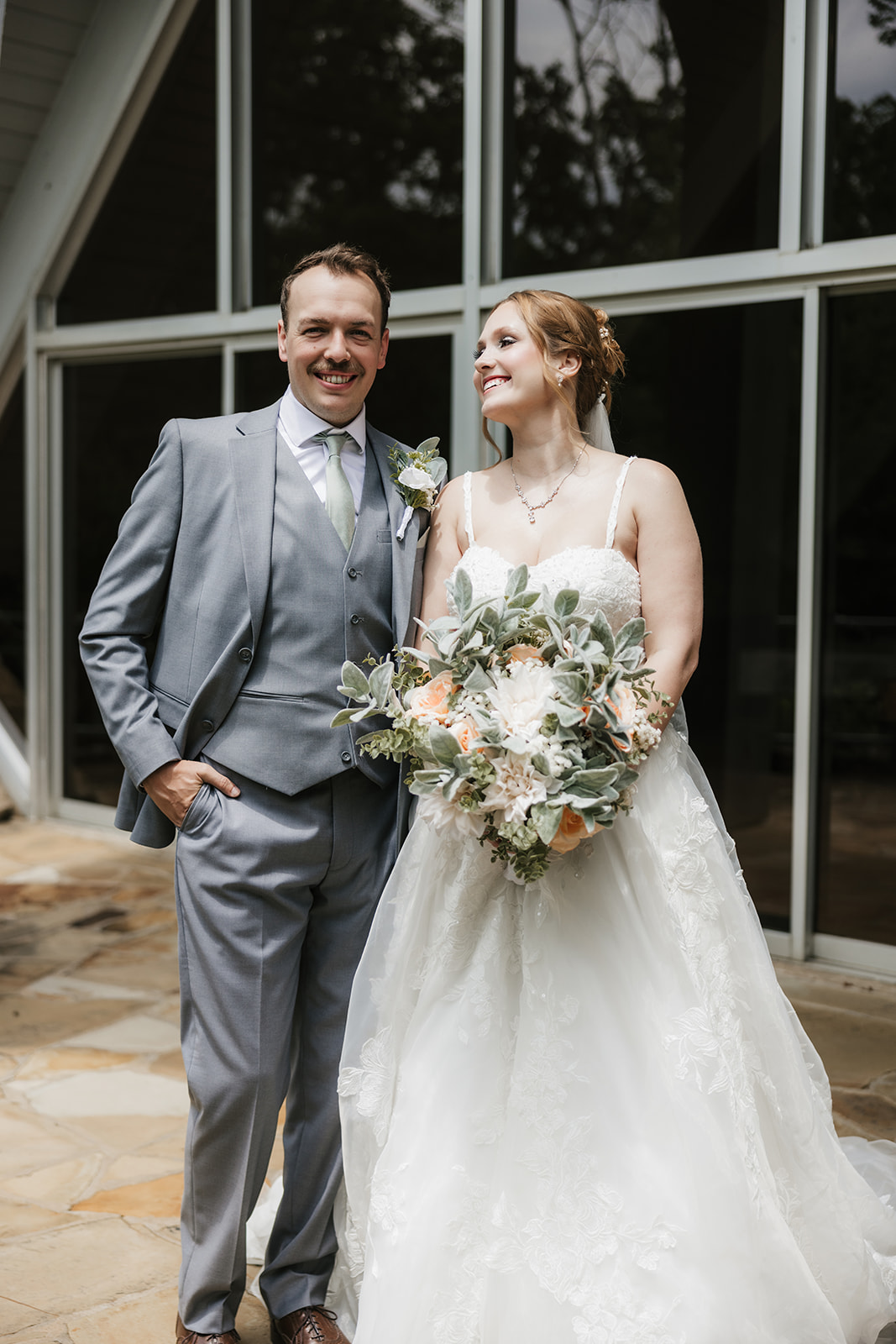 A bride and groom stand on a stone patio, sharing a kiss under the bride’s veil with trees and a metal railing in the background for a Camp Loughridge Wedding