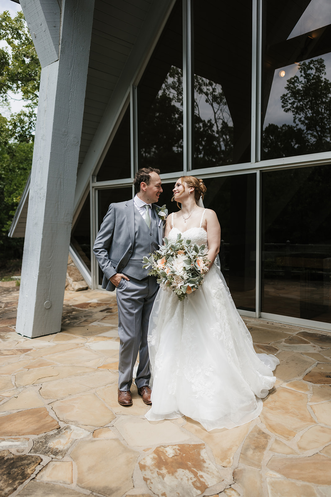 A bride and groom stand on a stone patio, sharing a kiss under the bride’s veil with trees and a metal railing in the background for a Camp Loughridge Wedding