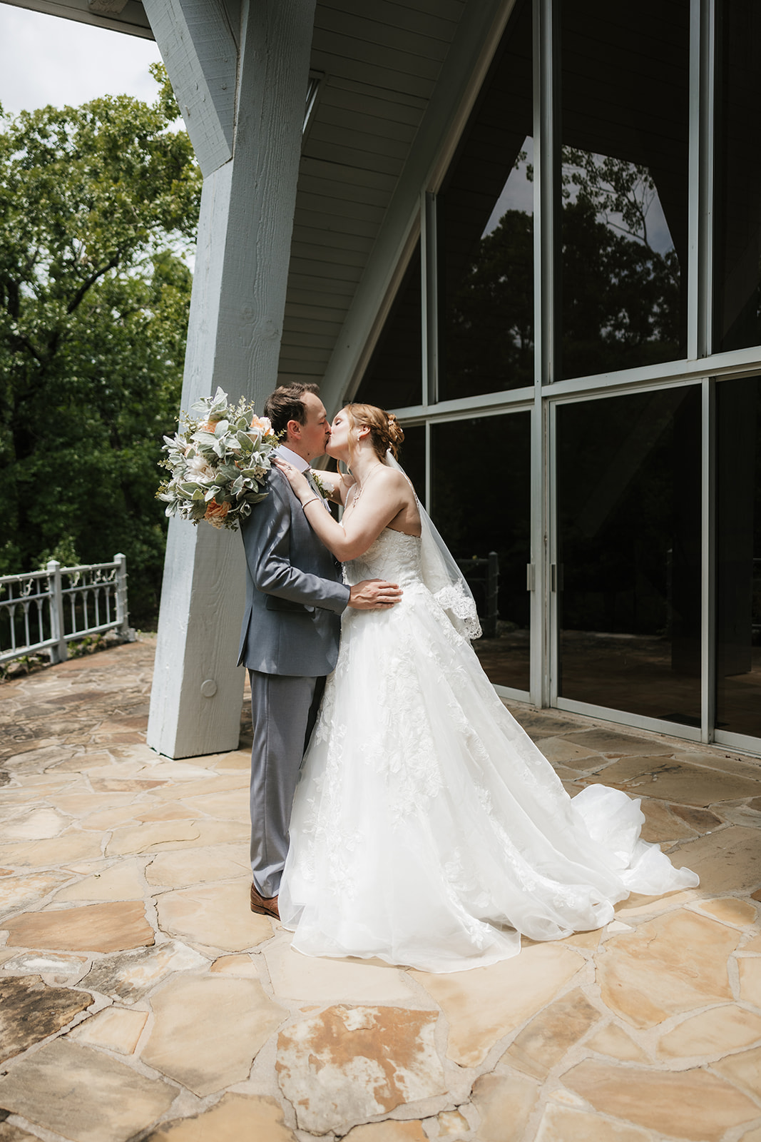 A bride and groom stand on a stone patio, sharing a kiss under the bride’s veil with trees and a metal railing in the background for a Camp Loughridge Wedding